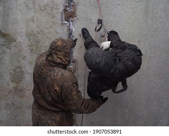 Training In Military, Assault And Police Mountaineering. Male Instructor Teaches A Man In Camouflage To Climb Down A Wall On A Rope. Special Forces Learning At The Training Ground. Photo Without Face.