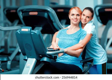 Training With Mom. Beautiful Mature Woman Bonding Her Teenage Daughter In Sports Clothing After Workout On Exercise Bicycle In The Gym