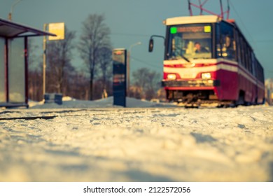 The Tram Arrives At The Stop. Sunrise On A Winter Morning. Snow-covered Road. Trees In The Park. Focus On Snow. Close Up View From Ground Level.