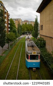 Tram Line Two In Göteborg, Sweden, Photographed From Behind When Its Approaching Tram Stop Brunnsgatan In Between Yellow Brick Buildings, Driving On Green Grass