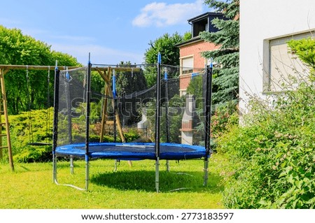 A trampoline is placed in a backyard surrounded by trees and a wooden swing set on a bright sunny day.