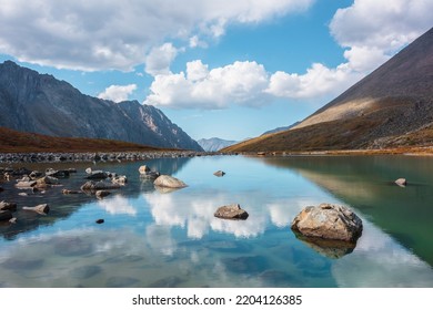 Tranquil autumn landscape with clouds reflection on smooth mirror surface of mountain lake in high hanging valley. Meditative view from calm alpine lake to mountain vastness. Stones in clear water. - Shutterstock ID 2204126385