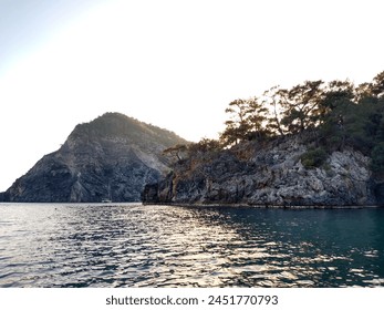 A tranquil bay at sunset. In the foreground there's a Grey and Orange rocky Cliff Face with Pine Trees covering it and a Yacht and Hill backdrop. The Sea is dark Jade and reflects the Sky light.   - Powered by Shutterstock