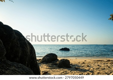 A tranquil beach landscape featuring smooth, golden sand and large, rugged rocks in the foreground. The calm ocean stretches into the horizon under a clear, blue sky, bathed in warm sunlight. Perfect for travel, relaxation, and coastal-themed projects. Ideal for stock use in advertising, tourism, or lifestyle content.