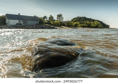 The tranquil Mērsrags beach in Latvia. The sunlit water shimmers around a large rock in the foreground, with a rustic building and lush greenery in the background, evoking a peaceful coastal scene. - Powered by Shutterstock