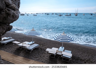 A tranquil beach on the Amalfi Coast showcases lounge chairs under striped umbrellas, with gentle waves lapping at the shore and boats dotting the horizon, inviting serenity and relaxation. - Powered by Shutterstock