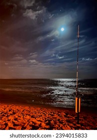 A tranquil beach scene with sand stretching along the shore, the moon reflecting on the calm sea, and a fishing rod casting its silhouette against the serene backdrop.