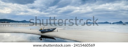 Tranquil beach scene with a solitary wooden boat on white sand, a long pier, and distant mountains under a cloudy sky