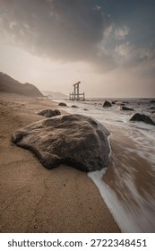 A tranquil beach scene at sunset, featuring a rocky shore with gentle waves lapping against the sand. A weathered wooden structure stands in the water, surrounded by large rocks. The sky is painted with hues of orange and blue, creating a peaceful atmosphere.
