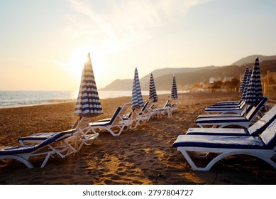 A tranquil beach scene at sunset featuring rows of blue-and-white striped umbrellas and matching lounge chairs along the sandy shore, inviting relaxation, warmth, and seaside leisure.