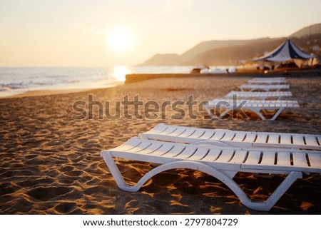 A tranquil beach scene at sunset with a row of white plastic lounge chairs on warm sand, gentle waves, and distant tents. Ideal for vacation, relaxation, travel, and holiday mood visuals.