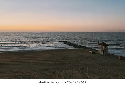  A tranquil beach scene at sunset, with soft hues of orange and pink lighting the horizon. Gentle waves roll onto the shore near a lifeguard tower and a rocky jetty stretching into the ocean - Powered by Shutterstock