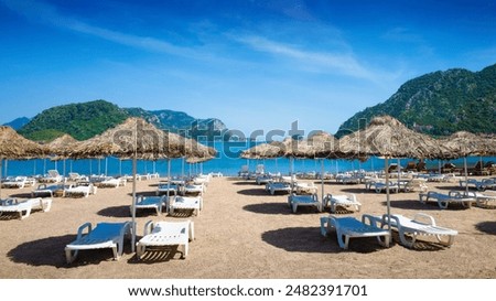 Tranquil beach setting in Marmaris with straw umbrellas, white sun loungers on sandy shore. Calm blue sea, picturesque mountains under clear sky. Welcome to Marmaris, Turkey concept image