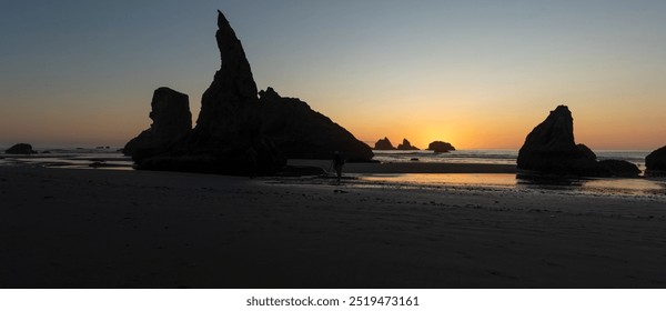 A tranquil beach at sunset with several large, jagged rock formations silhouetted against the orange and blue sky. The calm ocean waves gently wash up on the shore, reflecting the warm colors of the s - Powered by Shutterstock