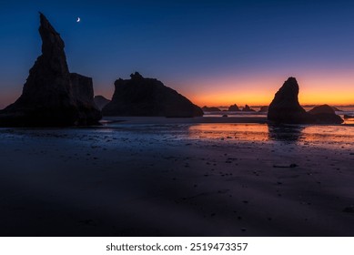 A tranquil beach at twilight features towering rock formations silhouetted against a colorful sky transitioning from deep blue to orange. A crescent moon hangs above, and the wet sand reflects the fad - Powered by Shutterstock