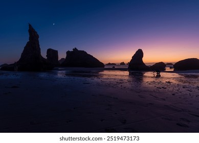 A tranquil beach at twilight with prominent rock formations silhouetted against a gradient sky transitioning from deep blue to warm orange. A crescent moon is visible above, and the wet sand reflects - Powered by Shutterstock