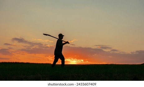 In the tranquil beauty of a rural evening, the silhouette of a genuine farmer emerges, walking with a shovel in the vast field. The dramatic sky sets the scene aglow with vibrant colors