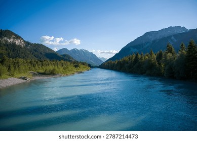A tranquil blue river winds through a forested valley between rugged mountains and snow-capped peaks, framed by evergreen trees and a clear sky for a peaceful alpine landscape scene.