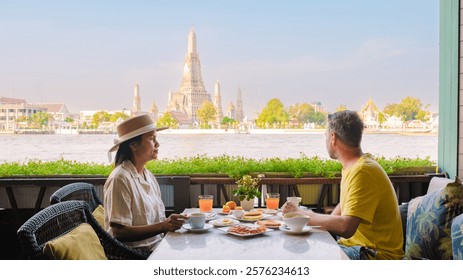 A tranquil breakfast setting on the river in Bangkok, where a couple savors delicious dishes while taking in a breathtaking view of the iconic Wat Arun temple and lush surroundings.