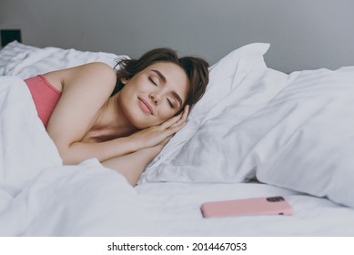 Tranquil calm young woman 20s in pajamas sleeping lying in bed wrap covered under blanket duvet on pillow near mobile cell phone rest relax indoors at home. Good mood night morning bedtime concept.