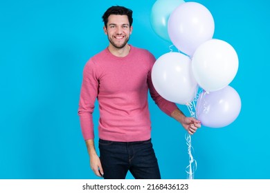 Tranquil Caucasian Guy Handsome Brunet Man With Bunch of Colorful Air Balloons in Pink Jumper Showing Smile Over Blue Background. Horizontal image Orientation