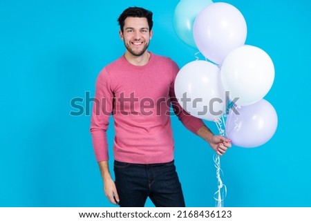 Tranquil Caucasian Guy Handsome Brunet Man With Bunch of Colorful Air Balloons in Pink Jumper Showing Smile Over Blue Background. Horizontal image Orientation