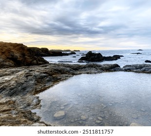 A tranquil coastal scene at dusk featuring rocky shores and tide pools under an overcast sky. The serene ocean view and rugged coastline create a peaceful atmosphere in this natural landscape. - Powered by Shutterstock