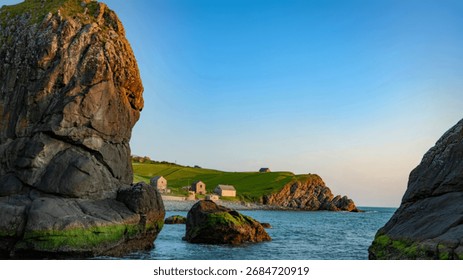 A tranquil coastal scene featuring dramatic, moss-covered cliffs framing a picturesque cove with several small stone buildings. The clear blue sky and calm sea enhance the serenity.