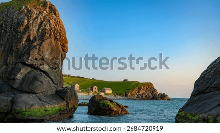 A tranquil coastal scene featuring dramatic, moss-covered cliffs framing a picturesque cove with several small stone buildings. The clear blue sky and calm sea enhance the serenity.