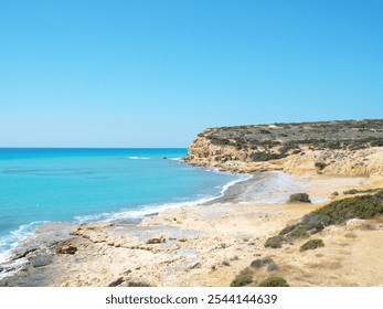 A tranquil coastal scene with turquoise waters, rocky beach, and rugged cliffs under a clear blue sky. The untouched natural landscape radiates calm and solitude. - Powered by Shutterstock