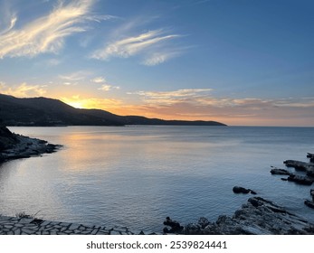 A tranquil coastal sunset with golden light reflecting on calm waters. The sky glows with soft clouds, and the rocky shoreline adds natural texture, evoking a peaceful evening by the sea.

 - Powered by Shutterstock