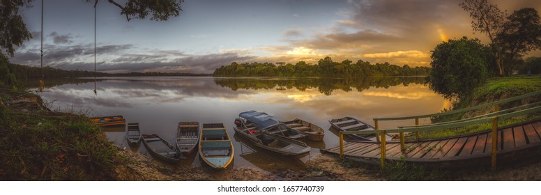Tranquil colorful orange tropical sunrise over a lake and moored boats with wooden walkway and reflections of the surrounding trees in a panorama banner view