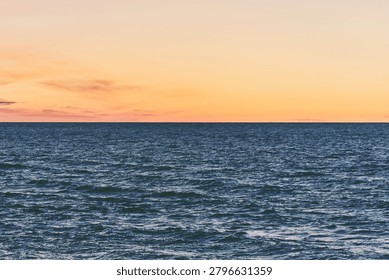Tranquil deep blue ocean surface stretches to crisp horizon against gentle orange and pink gradient sky at dusk, evoking peaceful and serene coastal atmosphere