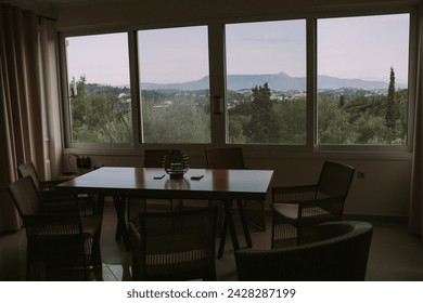 A tranquil dining room setting featuring a simple table arrangement and chairs, framed by large windows that offer a panoramic view of distant mountains and lush Mediterranean forestry.