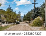 A tranquil dirt road in a small rural town, flanked by autumn trees and power lines, leading toward a scenic mountain backdrop.
