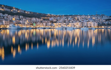 Tranquil evening scene of a charming coastal town with illuminated buildings and docked boats, beautifully reflected in the still harbor waters - Powered by Shutterstock