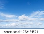 A tranquil and expansive view of a deep, clear blue sky filled with scattered, fluffy cumulus and wispy cirrus clouds, creating a peaceful scene above the calm blue waters of the sea horizon.