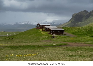 Tranquil Green Icelandic Landscape with Wooden Cabins and Snow-Capped Mountains in the Background Under a Moody Sky - Serene Nordic Rural Countryside Scene - Powered by Shutterstock