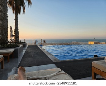 A tranquil infinity pool with ocean views at sunset, surrounded by palm trees and lounge chairs. A relaxing footrest moment captures the serene ambiance of the evening. - Powered by Shutterstock - Get 15% off with code: PIKWIZARD15