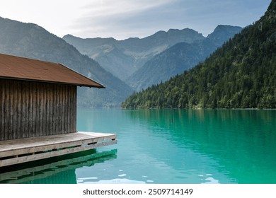 A tranquil lake nestled among majestic mountains showcases vibrant turquoise waters. A rustic wooden boathouse extends over the calm surface, inviting relaxation and reflection. Plansee Austria - Powered by Shutterstock