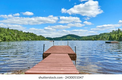 Tranquil lake reflects a vibrant blue sky dotted with fluffy clouds. Sunbeams dance on a long wooden pier stretching into the calm water. There are boaters and kayakers visible far in the lake - Powered by Shutterstock
