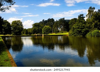 A tranquil lake set amidst lush greenery, capturing the calm reflections of trees and the clear blue sky above. The water's surface mirrors the serene environment, creating a peaceful and picturesque  - Powered by Shutterstock