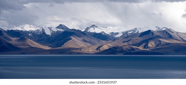 A tranquil lake sits in the foreground, with jagged, snow-capped mountains under a moody, overcast sky in the background. - Powered by Shutterstock