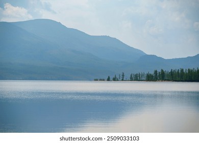 A tranquil lake with smooth waters reflects the soft light of a misty morning, framed by distant mountains and a tree-lined shore, creating a serene and peaceful landscape.  - Powered by Shutterstock