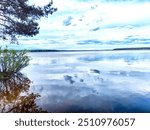 A tranquil lakeside scene with calm water reflecting clouds and trees under a partly cloudy sky.