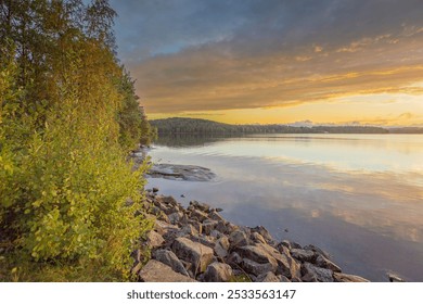 A tranquil lakeside scene with a rocky shore and lush greenery under a cloudy sunset sky. - Powered by Shutterstock