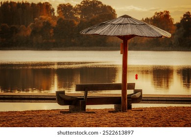 A tranquil lakeside scene at sunset, featuring a shaded bench under a beach umbrella. The golden glow reflects on the calm water, creating a peaceful atmosphere. - Powered by Shutterstock