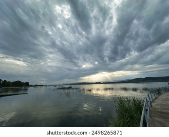 A tranquil lakeside scene under a dramatic, cloudy sky. Sunrays break through the clouds, casting light on the calm water, with boats docked along the shore and lush greenery lining the water's edge