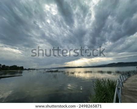 A tranquil lakeside scene under a dramatic, cloudy sky. Sunrays break through the clouds, casting light on the calm water, with boats docked along the shore and lush greenery lining the water's edge