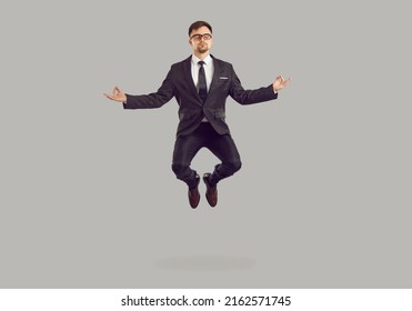 Tranquil Male Entrepreneur Levitating While Meditating. Studio Portrait Of Handsome Young Business Man In Suit And Glasses Practising Meditation Exercise, Reaching Zen And Floating In Air In Yoga Pose